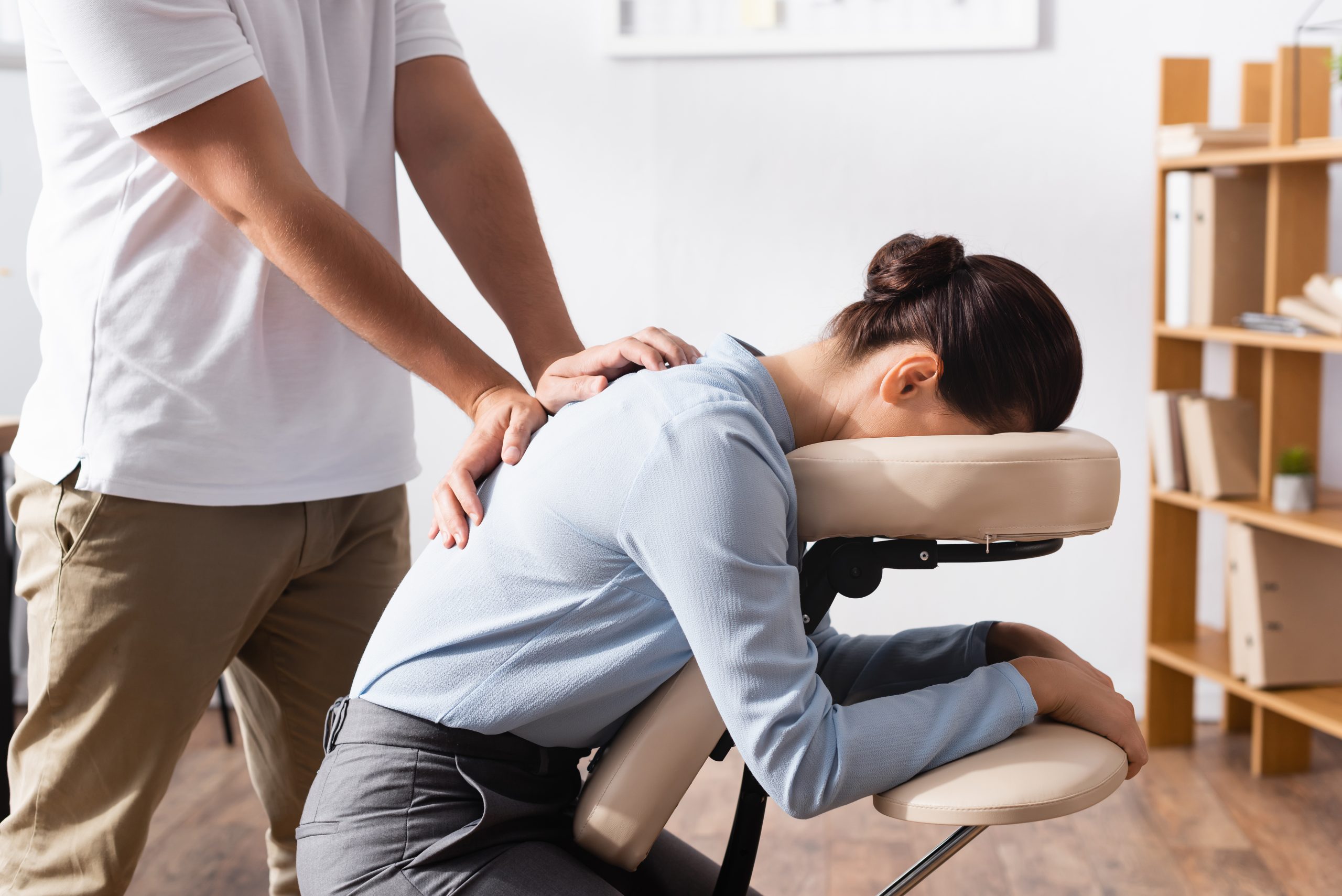 Side view of masseur doing seated massage of back for brunette businesswoman with blurred office on background