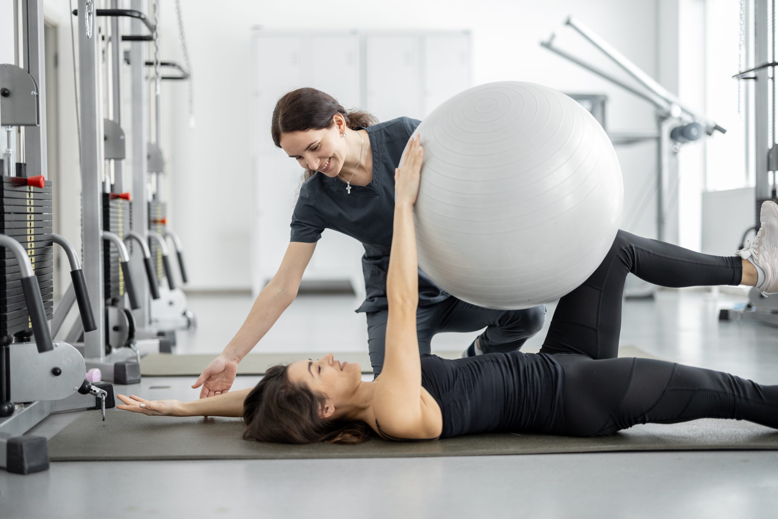 Woman doing exercises with fitness ball with rehabilitation specialist at the gym. Concept of physical therapy for health and recovery. Idea of recovery after pregnancy